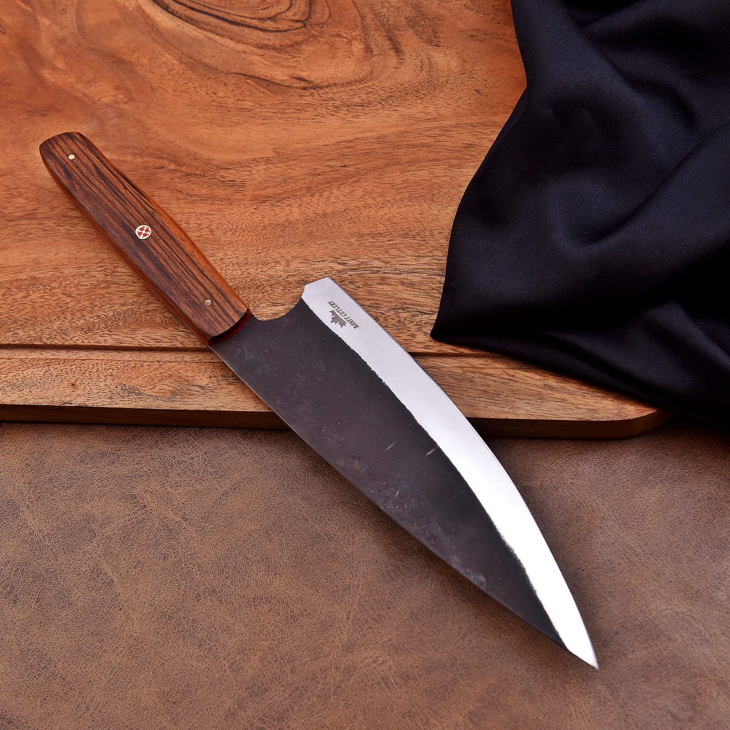 A handcrafted chef’s knife with a dark carbon steel blade and a polished wooden handle featuring a mosaic pin, resting on a wooden cutting board with a black cloth draped nearby.