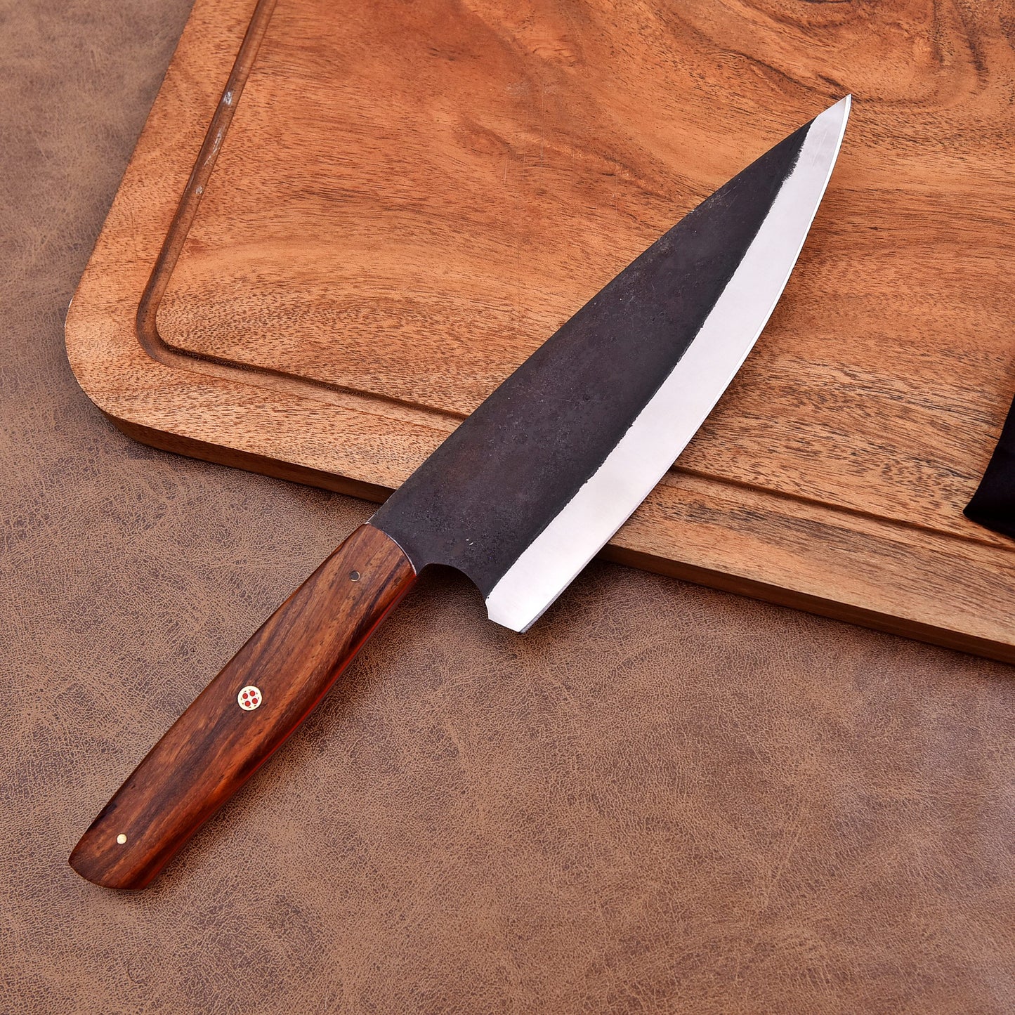 A handcrafted chef's knife with a dark, forged carbon steel blade and a polished wooden handle featuring a mosaic pin, displayed on a leather surface beside a wooden cutting board.