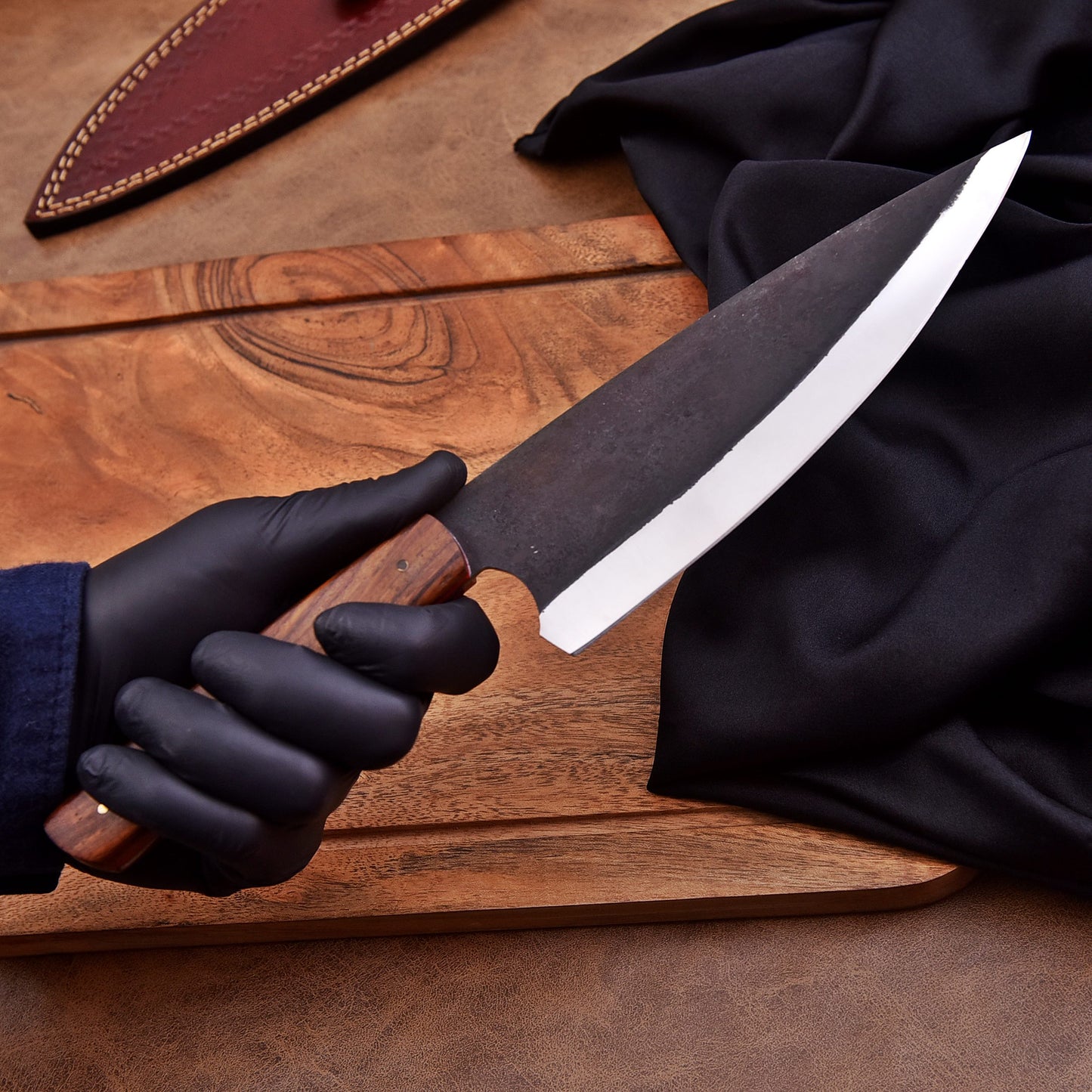 A person wearing black gloves holding a handcrafted chef’s knife with a dark carbon steel blade and wooden handle, positioned over a wooden cutting board with a black cloth and leather sheath nearby.