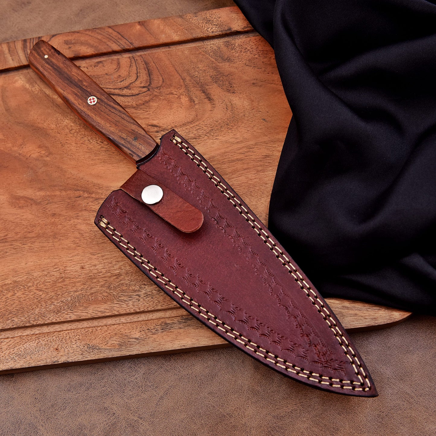 A handcrafted chef's knife partially sheathed in a brown leather case with decorative stitching and snap closure, resting on a wooden cutting board beside a black cloth.