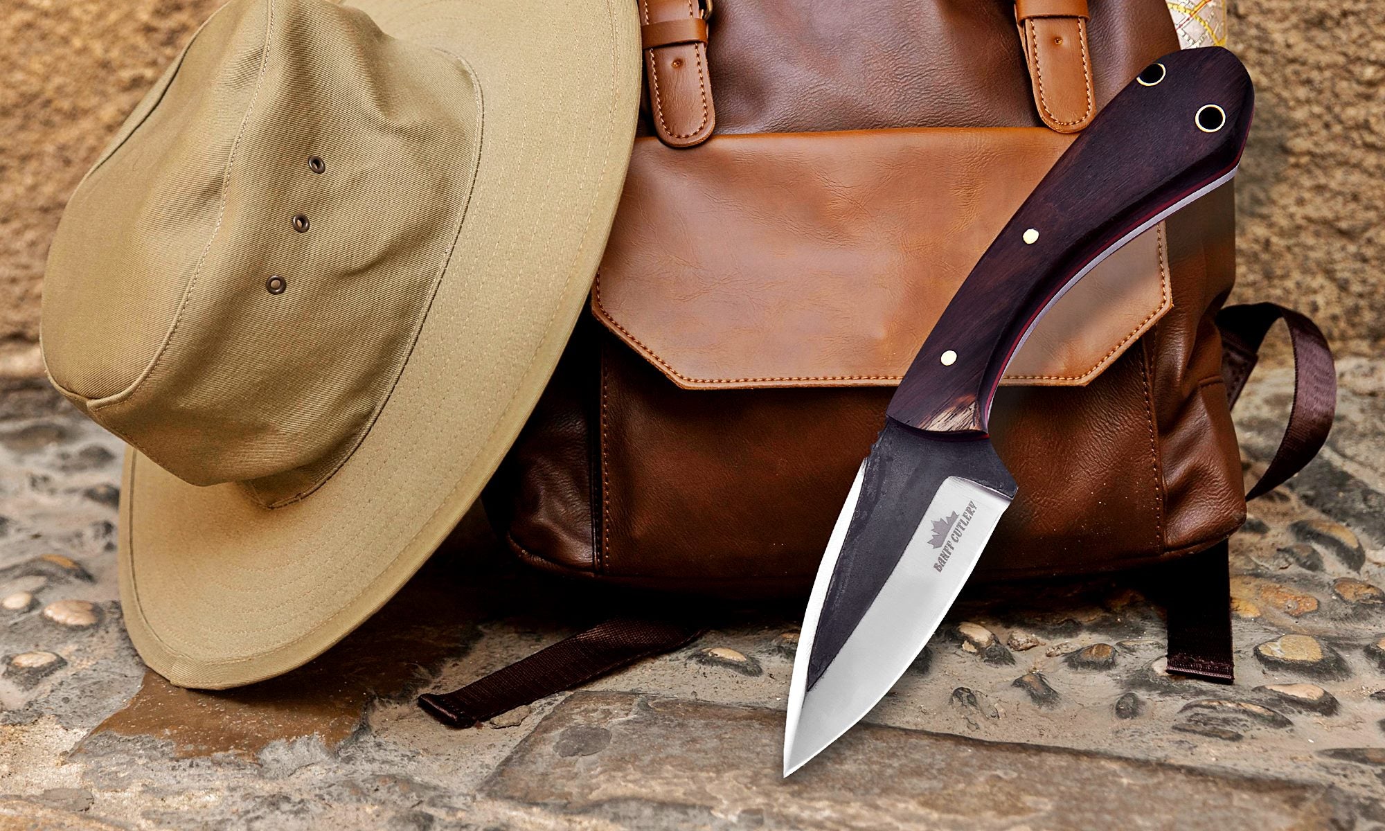 Fixed blade cowboy knife with wooden handle resting on leather backpack and safari hat – premium outdoor survival and Western-style knife gear