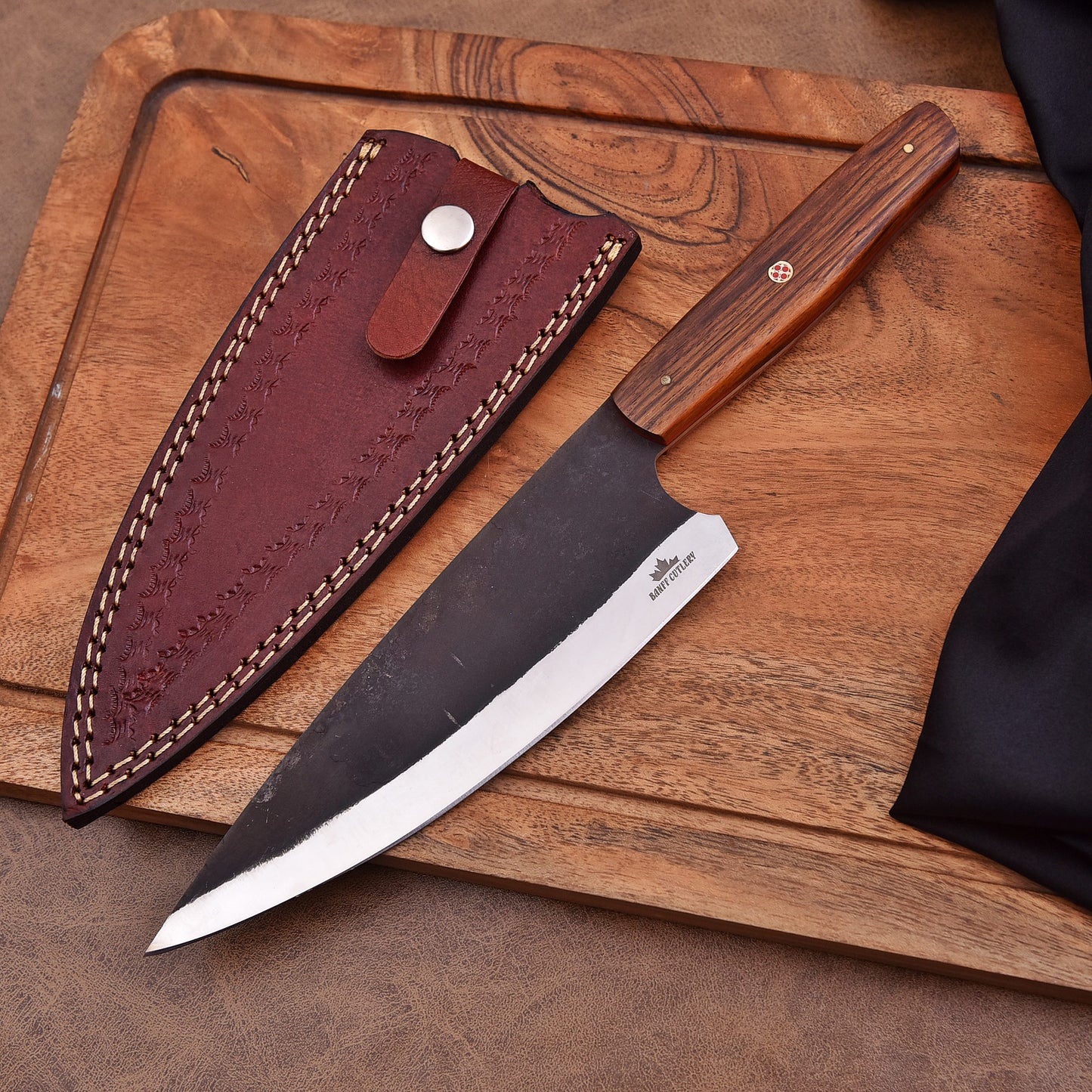 A handcrafted chef’s knife with a dark carbon steel blade and a wooden handle featuring a mosaic pin, displayed on a wooden cutting board beside a brown leather sheath with decorative stitching and a snap closure.