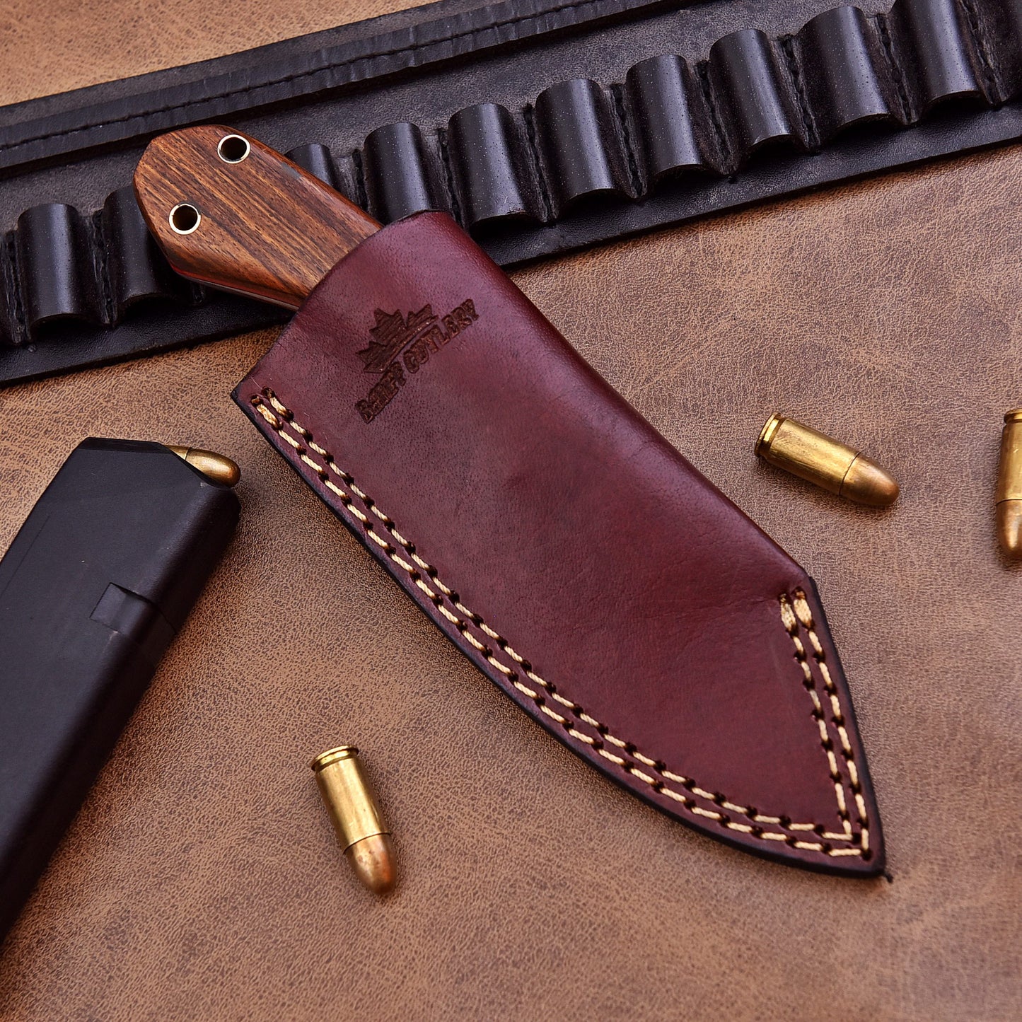A fixed-blade knife partially sheathed in a dark brown leather case embossed with the Banff Cutlery logo, lying on a leather surface alongside a black magazine and scattered bullets, with a black leather ammo belt in the background.