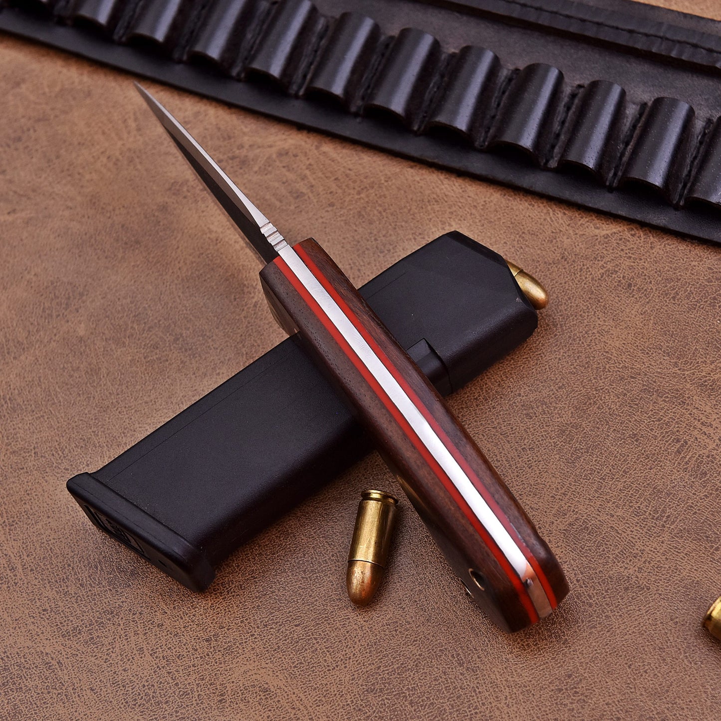 A side view of a fixed-blade knife showing the spine of the blade and the full tang construction with red liners and polished wooden handle scales. The knife is placed on top of a black magazine with scattered bullets and a leather ammo belt in the background, all set on a brown leather surface.