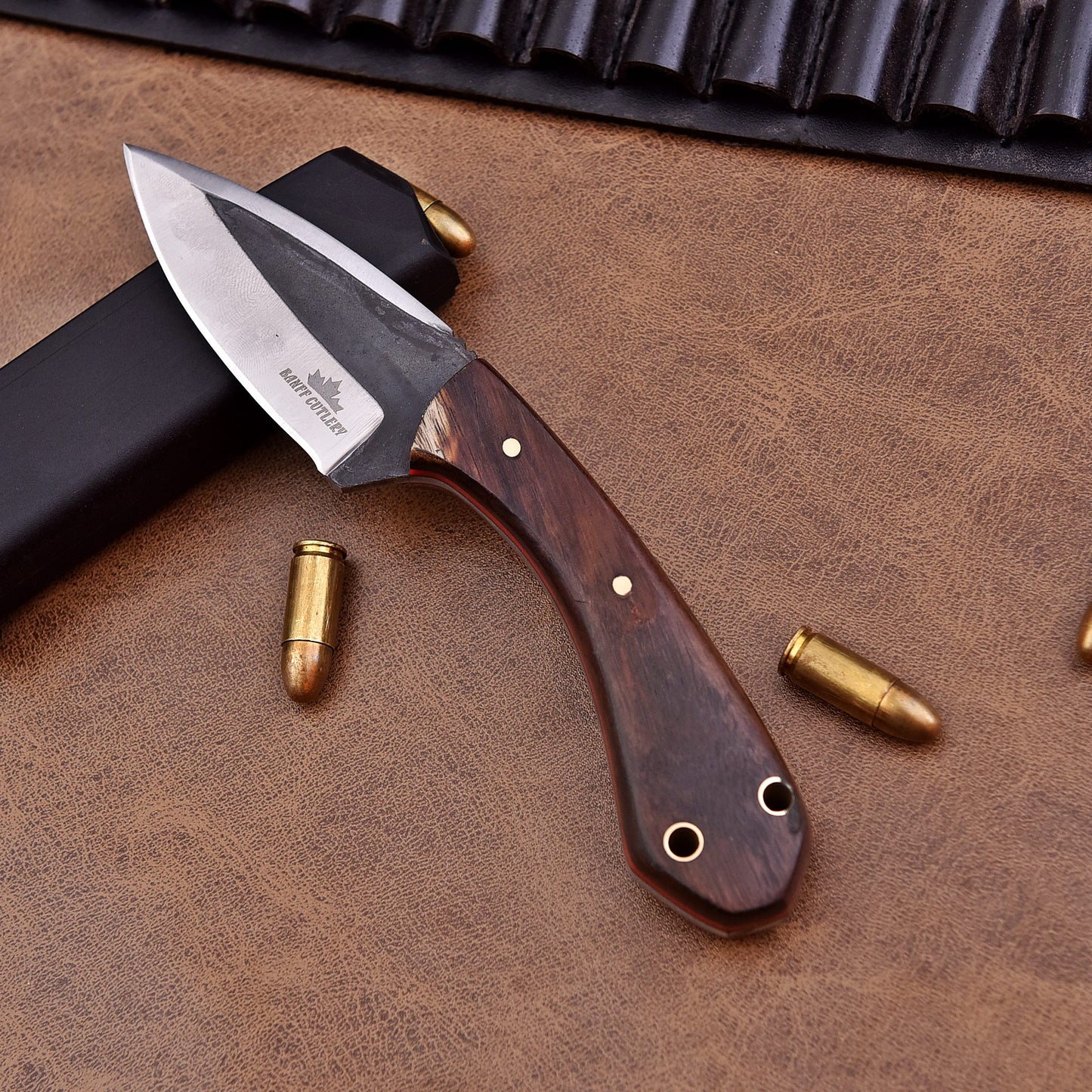 A fixed-blade knife with a polished wooden handle and a two-tone stainless steel blade engraved with the Banff Cutlery logo, placed on a leather surface beside scattered bullets, a black magazine, and a leather ammunition belt.