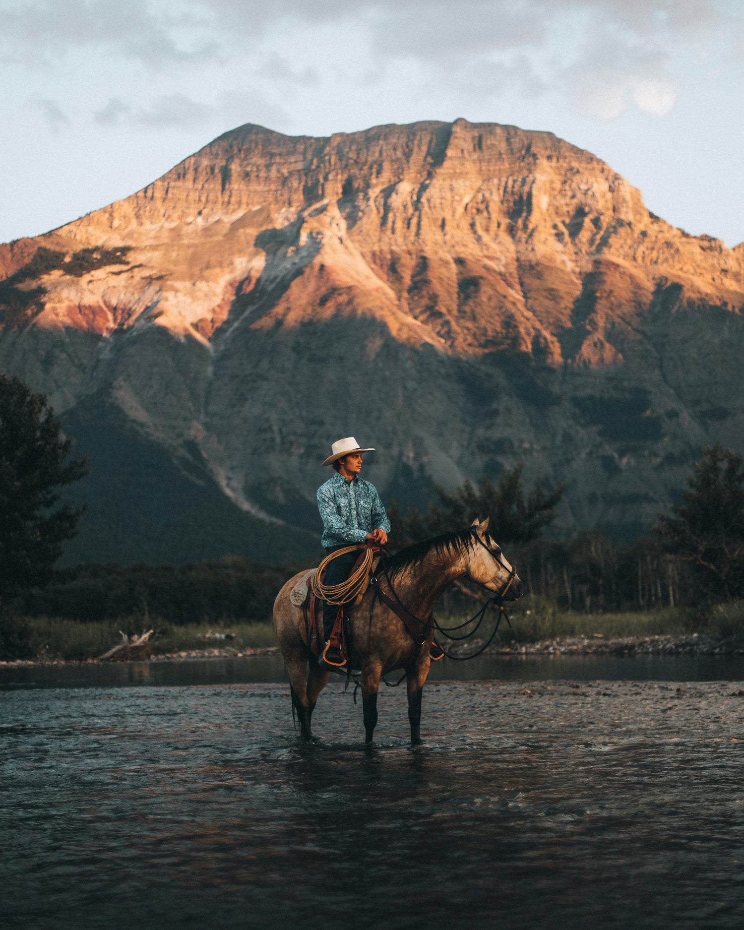 Cowboy on horseback crossing a river with a scenic mountain backdrop at sunset – Western lifestyle and rugged outdoor adventure imagery.