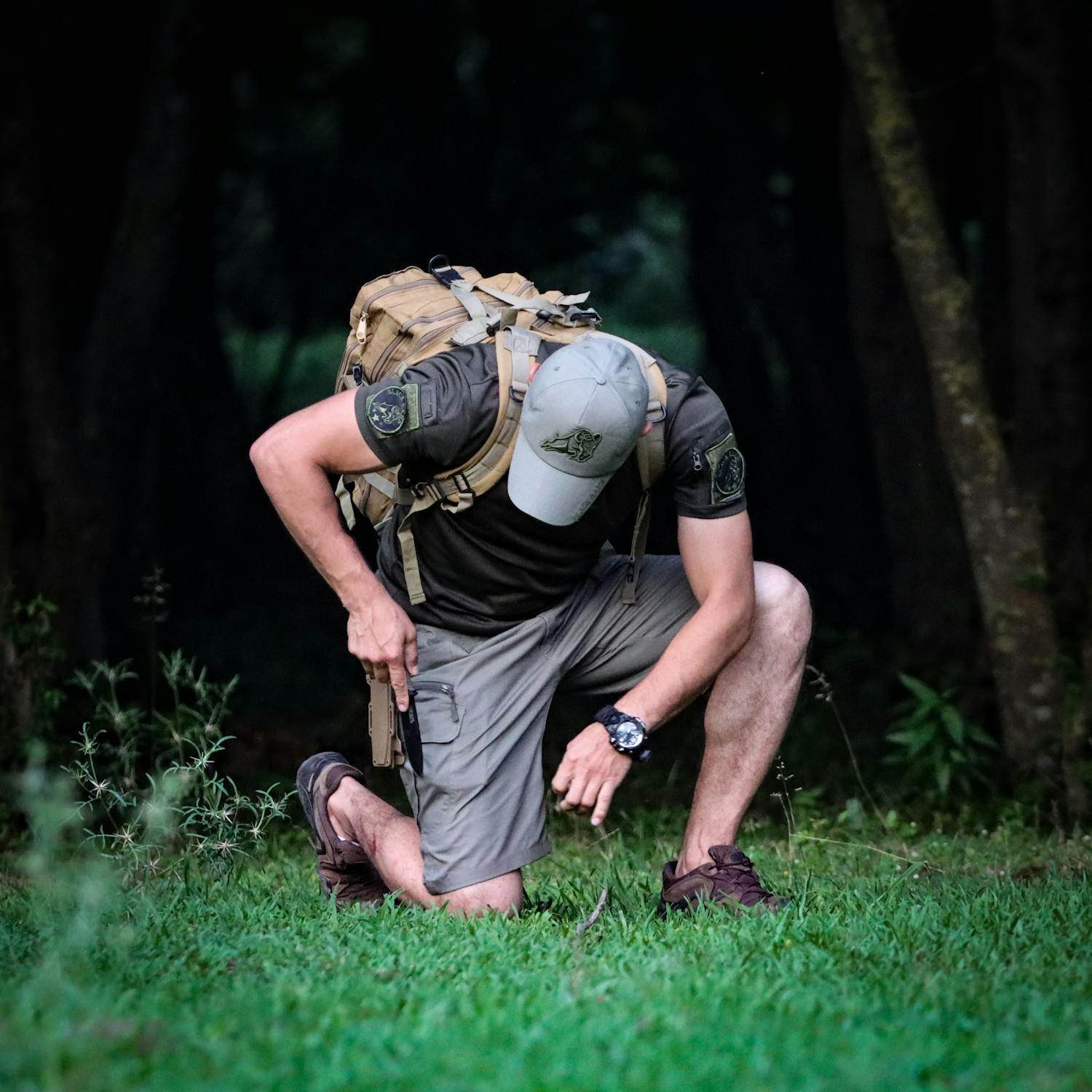 Man in tactical gear crouching in forest with backpack and pistol, dressed in outdoor survival clothing and camouflage hat, practicing wilderness preparedness or tactical training