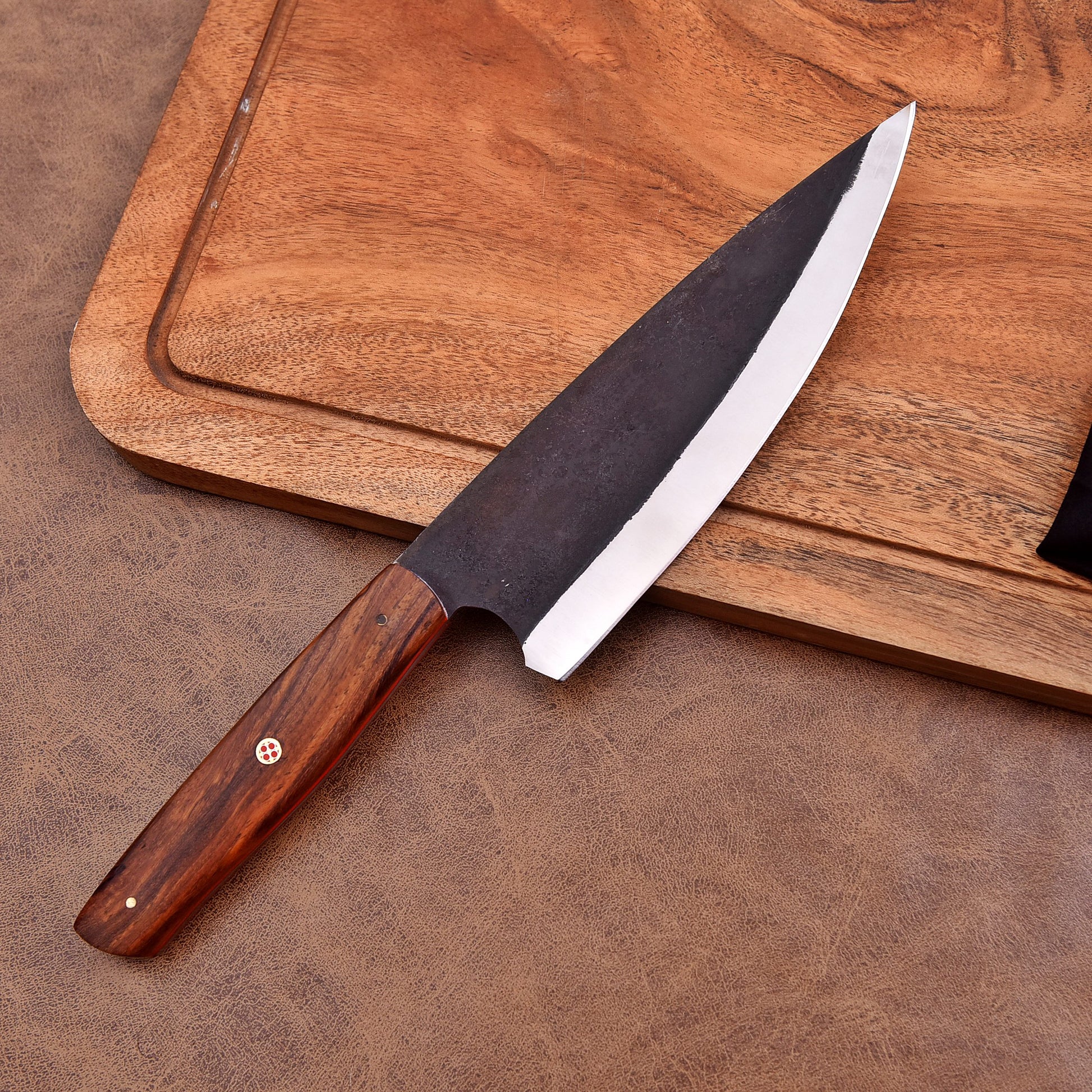 A handcrafted chef's knife with a dark, forged carbon steel blade and a polished wooden handle featuring a mosaic pin, displayed on a leather surface beside a wooden cutting board.