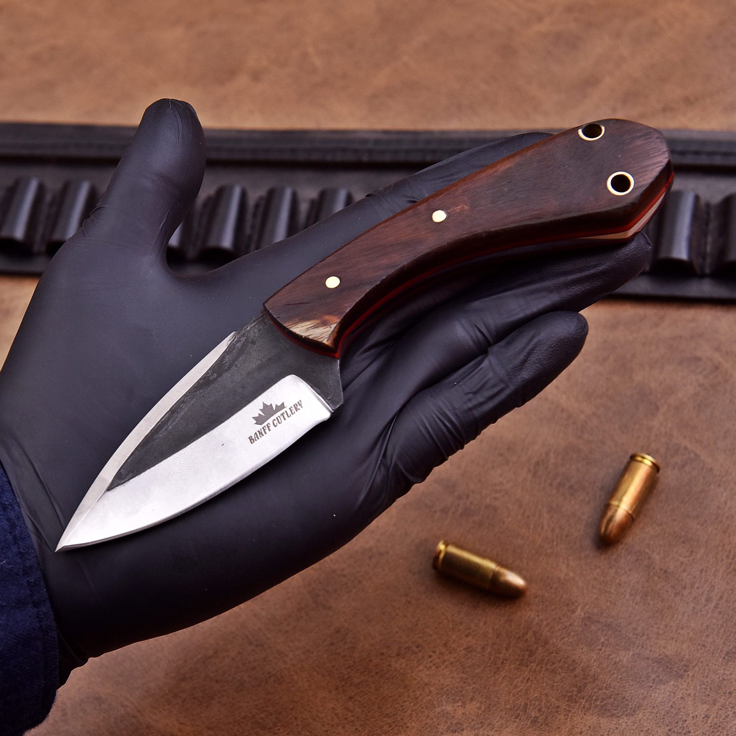 A gloved hand holding a fixed-blade knife with a polished wooden handle and a two-tone stainless steel blade engraved with the Banff Cutlery logo. The background features a leather surface with scattered bullets and a black leather ammo belt.