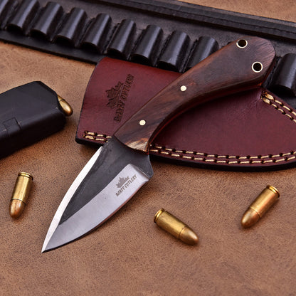 A fixed-blade knife with a polished wooden handle and stainless steel blade, engraved with the Banff Cutlery logo, resting on a leather sheath. The setup includes scattered bullets, a magazine, and a leather cartridge belt, all placed on a brown leather surface.