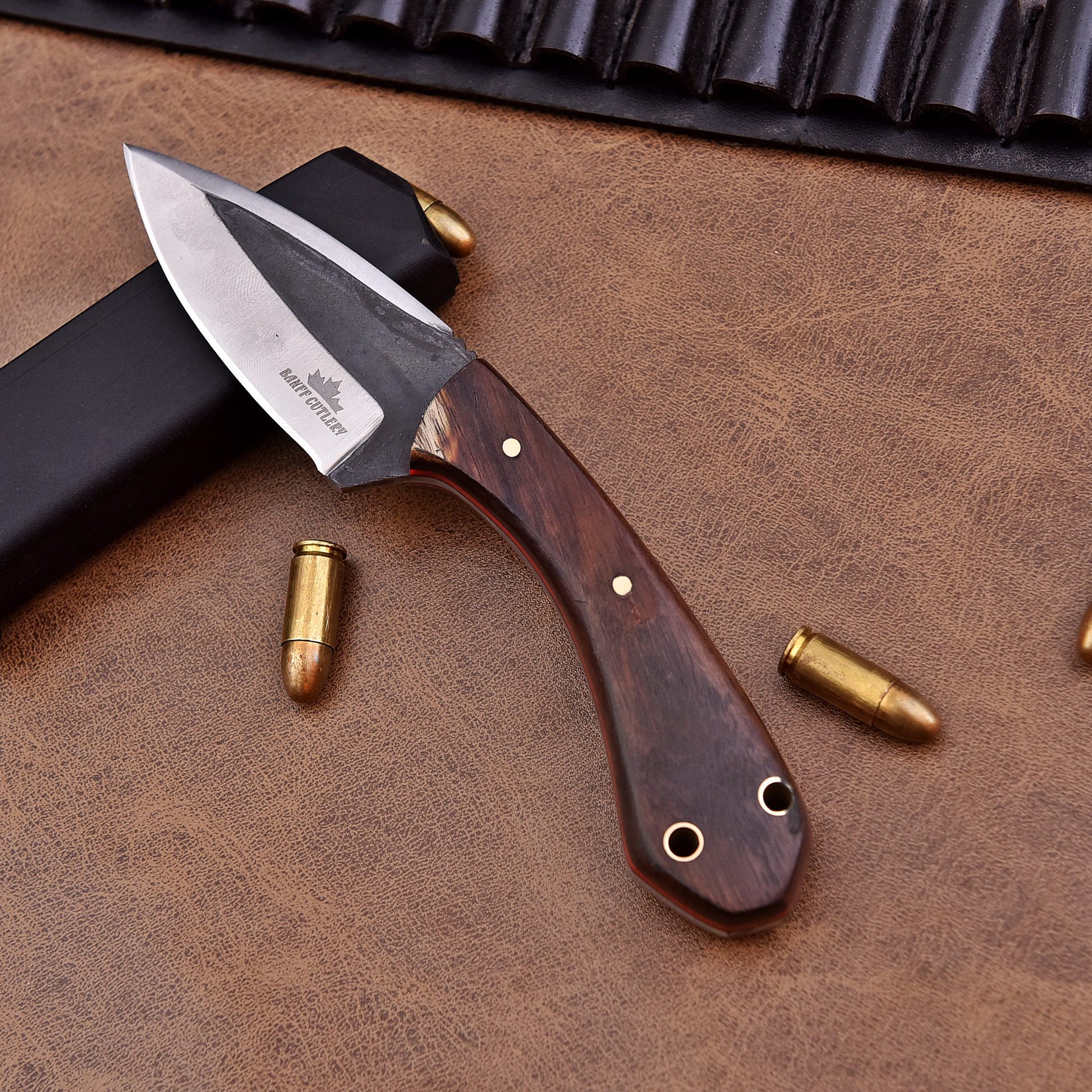 A fixed-blade knife with a polished wooden handle and a two-tone stainless steel blade engraved with the Banff Cutlery logo, placed on a leather surface beside scattered bullets, a black magazine, and a leather ammunition belt.