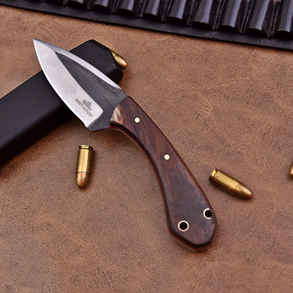 A fixed-blade knife with a polished wooden handle and a two-tone stainless steel blade engraved with the Banff Cutlery logo, placed on a leather surface beside scattered bullets, a black magazine, and a leather ammunition belt.